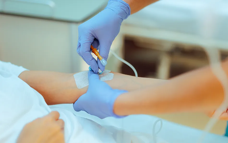 Zwei Hände in Handschuhen an einer Venenkanüle in der linken Armbeuge