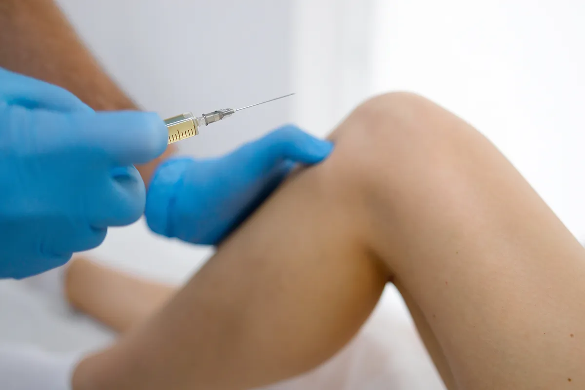 Eine Spritze in der Hand eines medizinischen Fachpersonals vor einem Knie