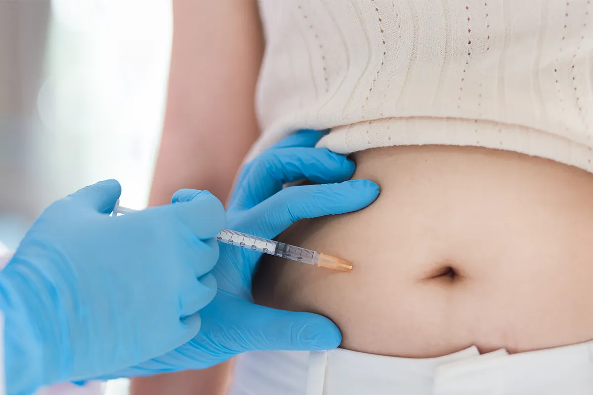 Eine Spritze in der Hand einer Person. Die Spritze steckt im Bauch einer weiblichen Person