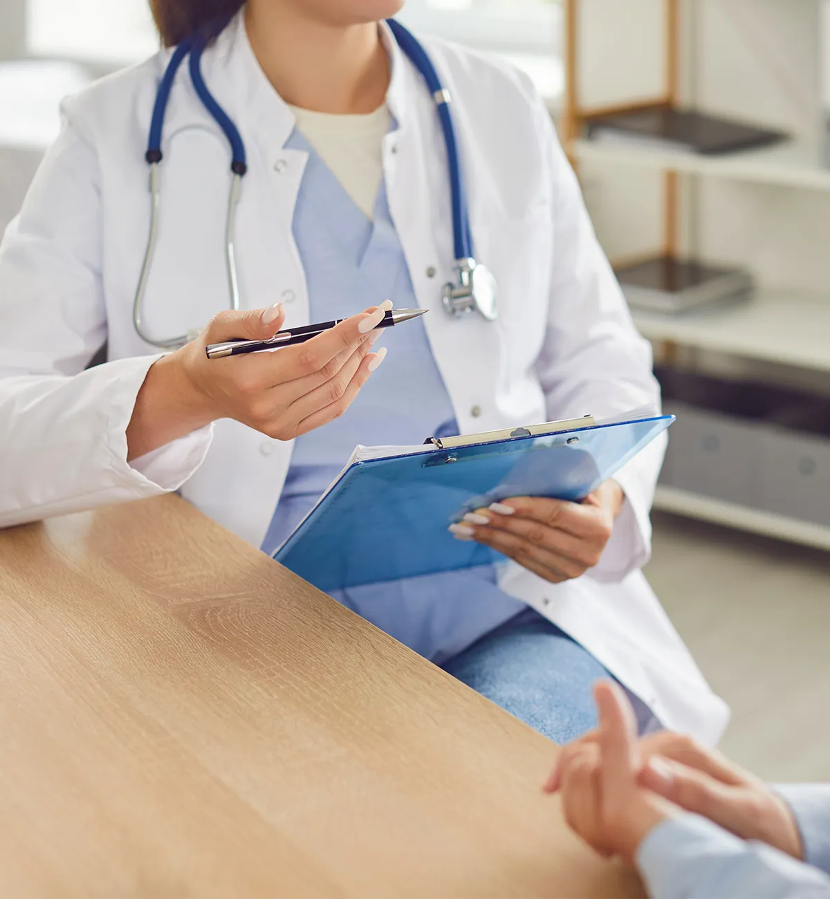 Medizinisches Personal sitzend mit Stetoskop um den Hals, sowie Aktenhalter und Stift in der Hand vor einer anderen Person an einem Tisch mit Regal im Hintergrund