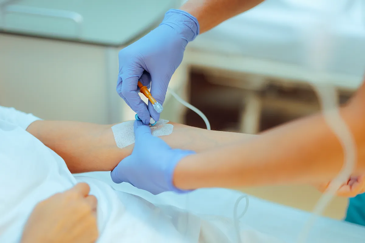 Zwei Hände in Handschuhen an einer Venenkanüle in der linken Armbeuge