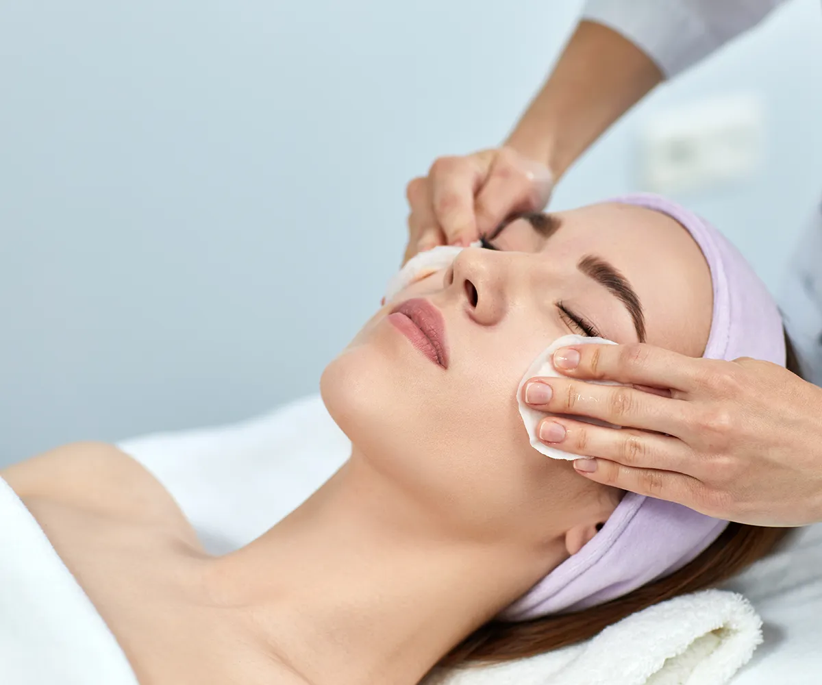 Lidogalen Creme vor Anwendung Unterspritzungen Eine Person wischt das Gesicht einer liegenden weiblichen Person mit Wattepads ab