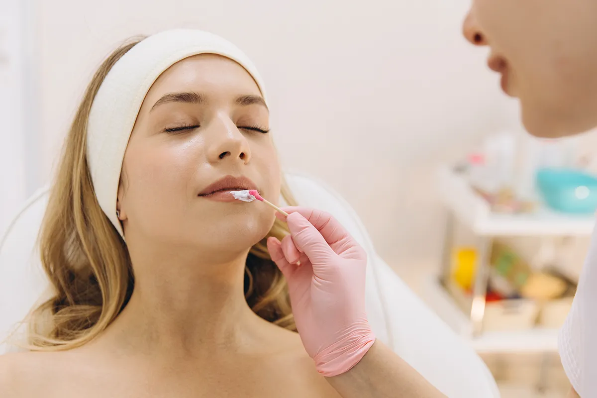 Einer weiblichen Person mit geschlossenen Augen wird eine Creme auf die Lippen aufgetragen